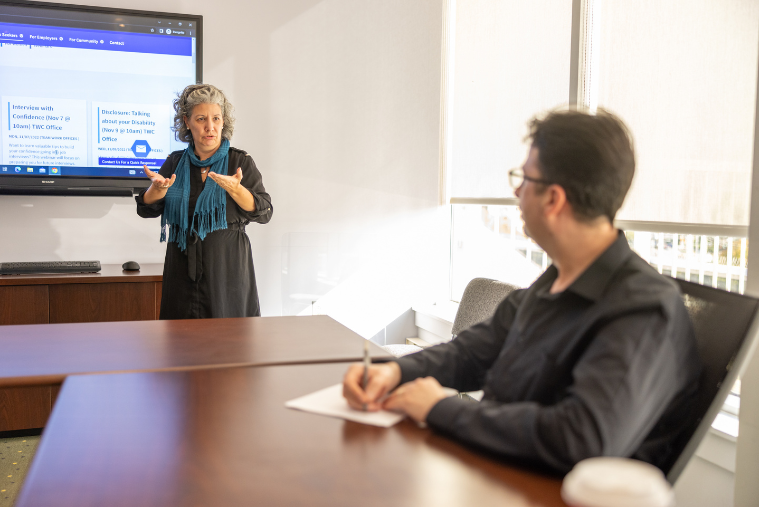 Woman presenting on a screen with a man blurred out in the foreground taking notes