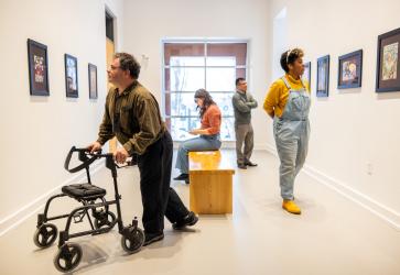 6321 - Group in a small gallery looking at paintings. One light-skinned man is using a walker, another light-skinned person is sitting on a bench in the centre of the gallery, and a medium-skinned person and dark-skinned person stand observing paintings on the opposite wall.