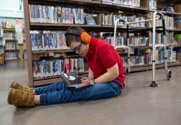 7781 - A light-skinned teenager with a visible disability wearing orange ear defenders sits on the floor hunched over outstretched legs looking at two DVDs in their lap. They are in a library and there is a walker behind them. 