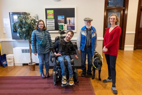  5061 - Group shot of four light-skinned people including one who is using a power wheelchair and another elder person who has their eyes closed and is holding onto a guide dog in harness. Two have beards and the other two have longer hair. 