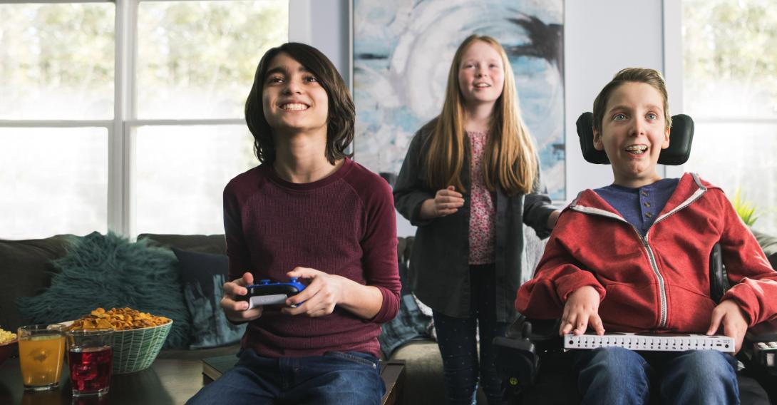Three smiling children playing a video game, the boy on the right uses a wheelchair