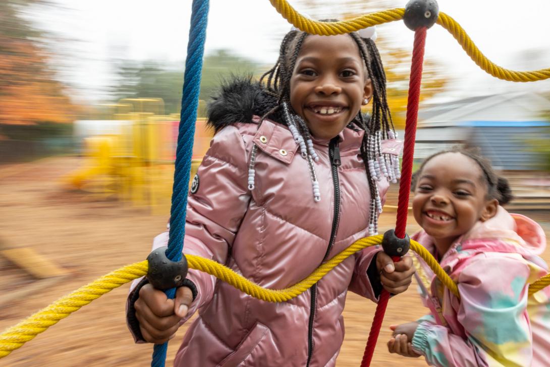 0141 - A smiling young Black girl with beaded braids and pink winter jacket holds onto a spinning rope play structure. Another younger Black girl hangs on beside her with a toothy grin. They are frozen in motion. 