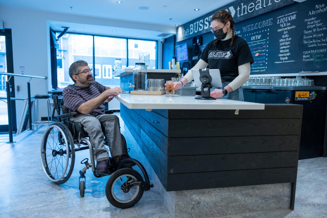 0448 - A customer with a visible physical disability using a manual wheelchair is paying a cashier for a drink over a lowered counter at a bar. 