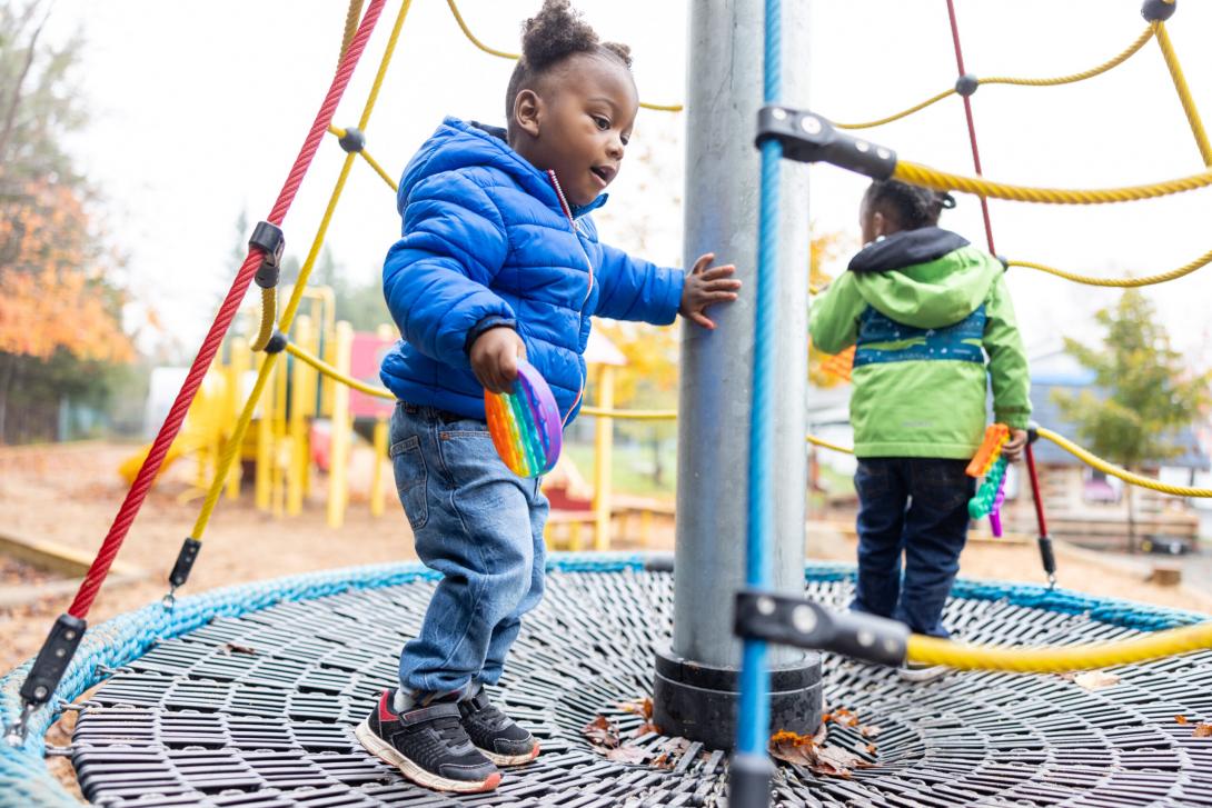 1917 - Two Black toddlers standing on a rope play structure, each holding a fidget toy and wearing fall jackets. The toddler in front has a steadying hand on the pillar at the centre of the structure.