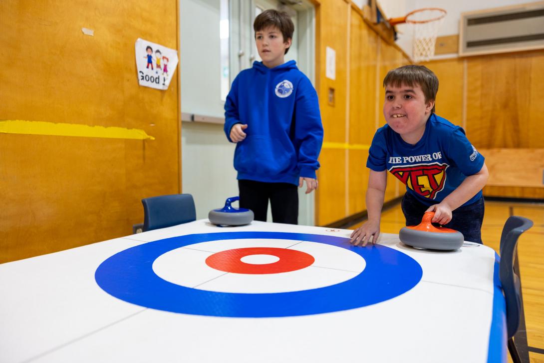 3459 - Two light-skinned children playing adapted curling on a raised inflatable shuffleboard in a school gym. One has a visible physical disability and is leaning over about to push the stone in their hand forwards. 