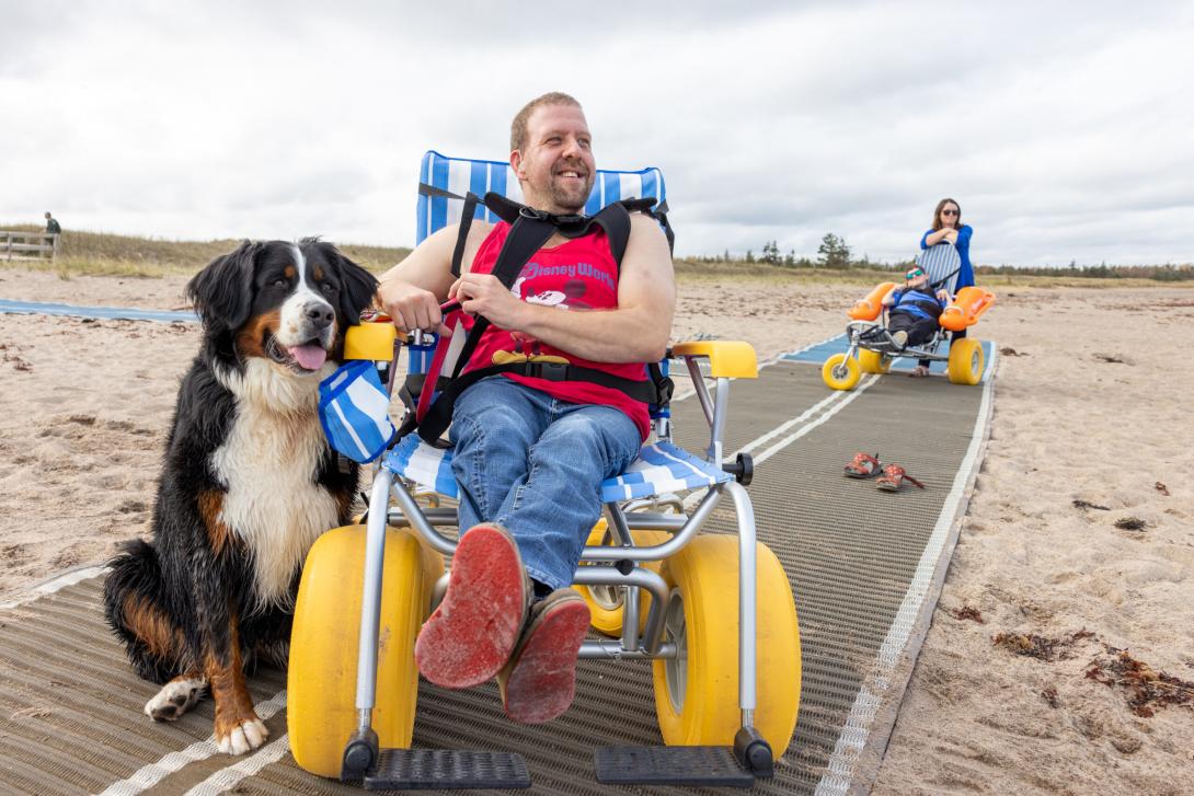 5427 - Adult relaxing in a floatable beach wheelchair on a mobility mat laid on a sandy beach beside a Bernese Mountain dog. They have light skin and facial hair. Two people including another in a floatable beach wheelchair are further behind them on the mobility mat. 