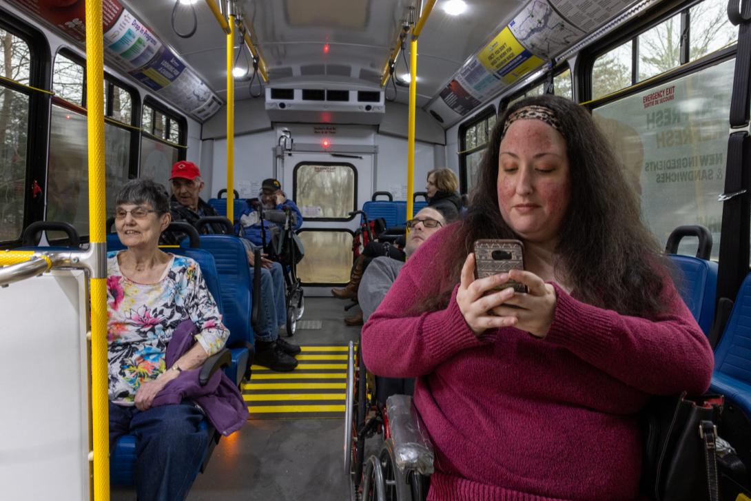 Passengers inside a bus including an elderly light-skinned woman, a younger light-skinned adult woman seated in a wheelchair in the front using their phone, and another wheelchair user behind them. The bus has yellow indicator strips on the ramp inside and vertical yellow poles. 