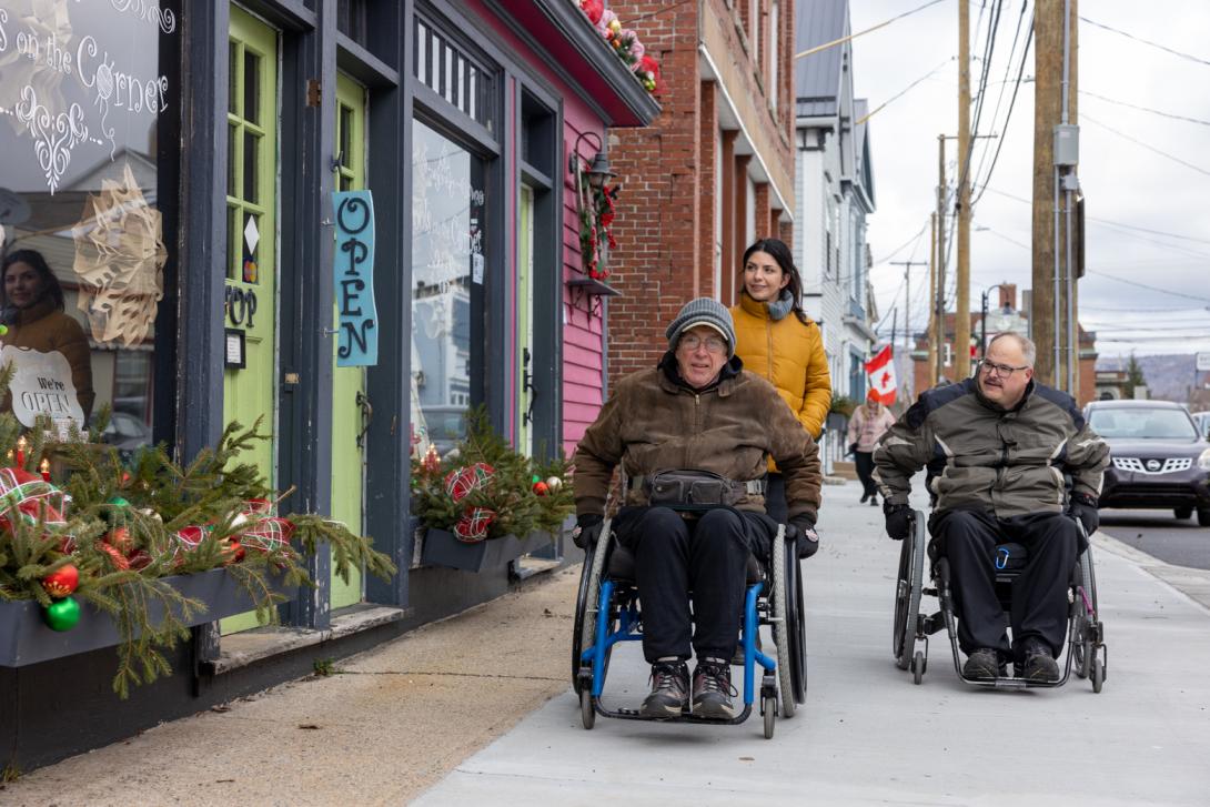 7689 - Group moving down a broad main street sidewalk including two light-skinned, male wheelchair users in warm jackets and a woman walking behind them. Colourful storefronts have outdoor Christmas decorations. 