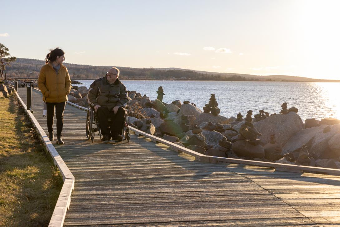 Two people conversing on a boardwalk at sunset. One is using a wheelchair and the other is walking. The coastline between the boardwalk and water is filled with piles of boulders, some with small inukshuks on them. 