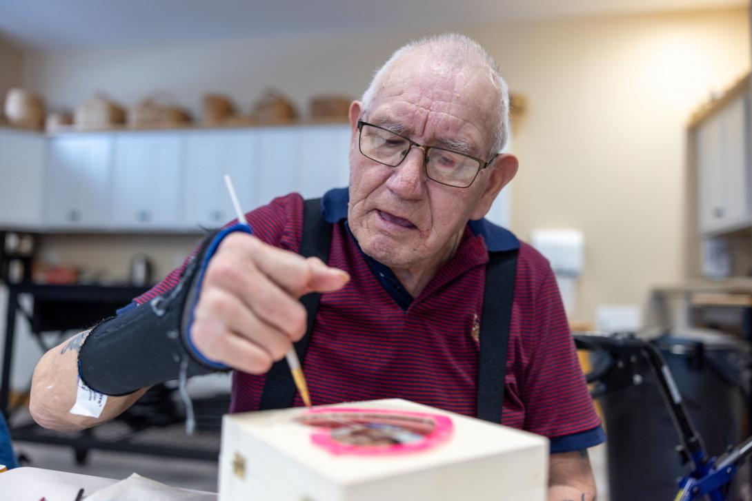 An elderly light-skinned man concentrates while painting a pink heart on a wooden box. He is wearing glasses and wrist brace and the handles of a walker are visible behind him.  
