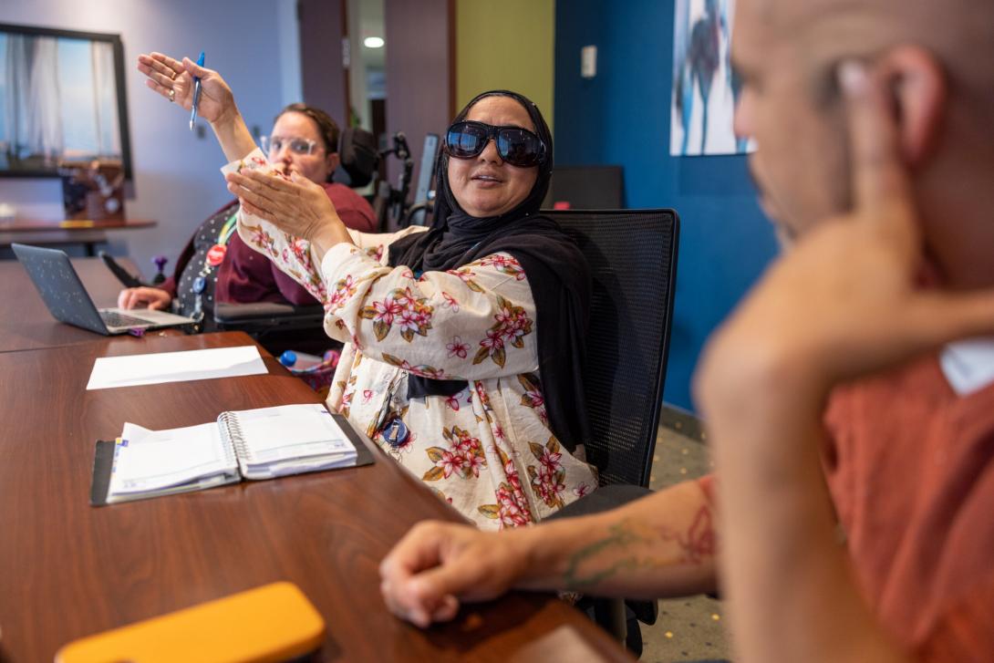 8846 - Three people seated in a board room meeting with laptops and notebooks. A power wheelchair user sits beside a person wearing dark glasses and a hijab who is speaking animatedly and gesturing. On the other end of the table is a person listening carefully to the person speaking.