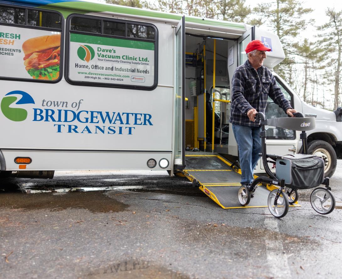 Adult wearing a red ball cap, plaid shirt and jeans using a walker to exit a Town of Bridgewater Transit bus in a parking lot.