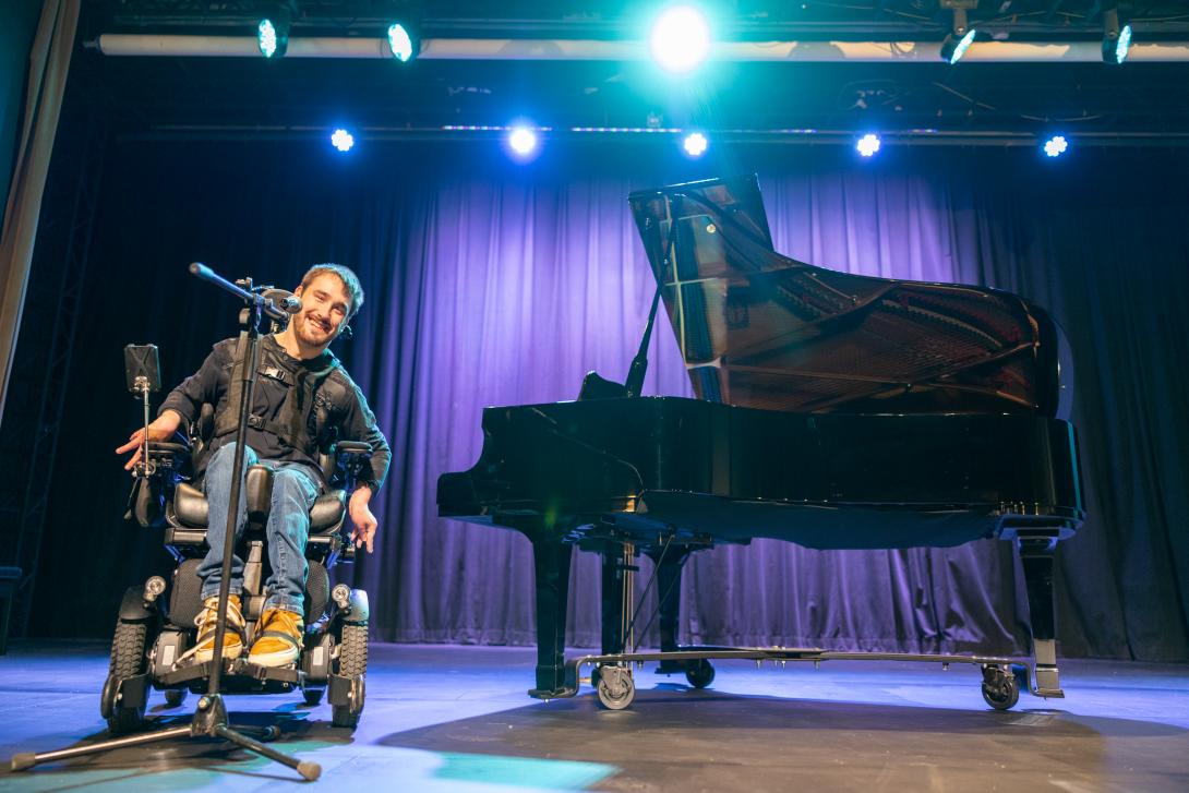 Man in wheelchair on stage next to a piano speaking into microphone
